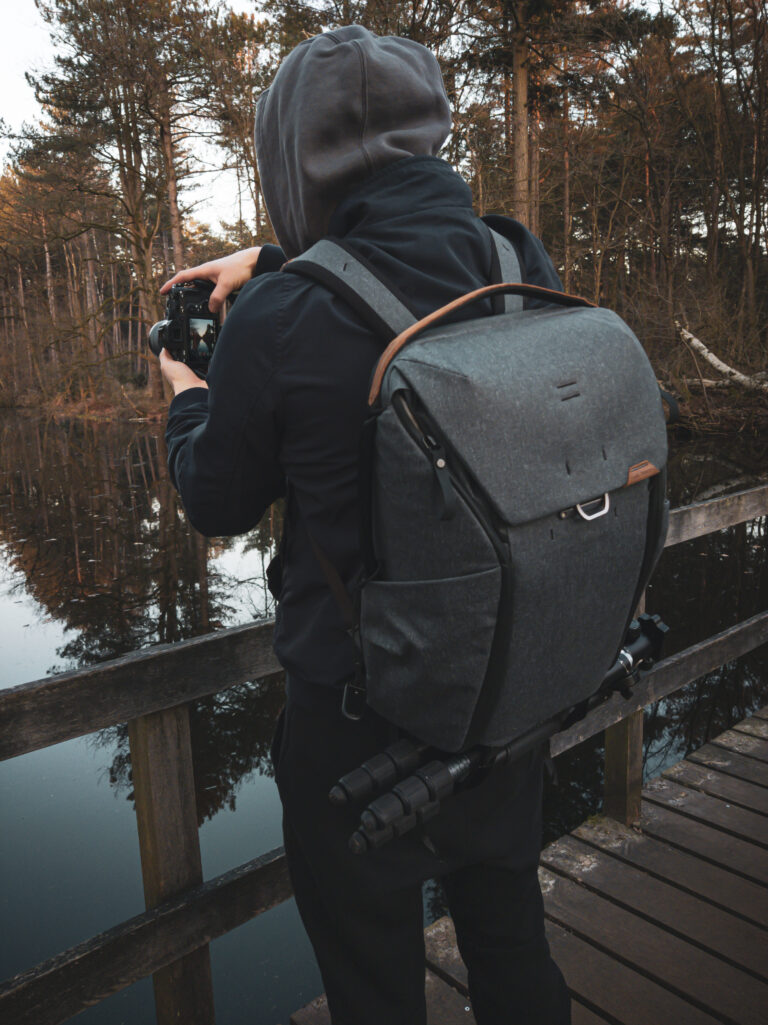 Szary plecak Everyday Backpack na plecach osoby fotografującej przy drewnianej barierce