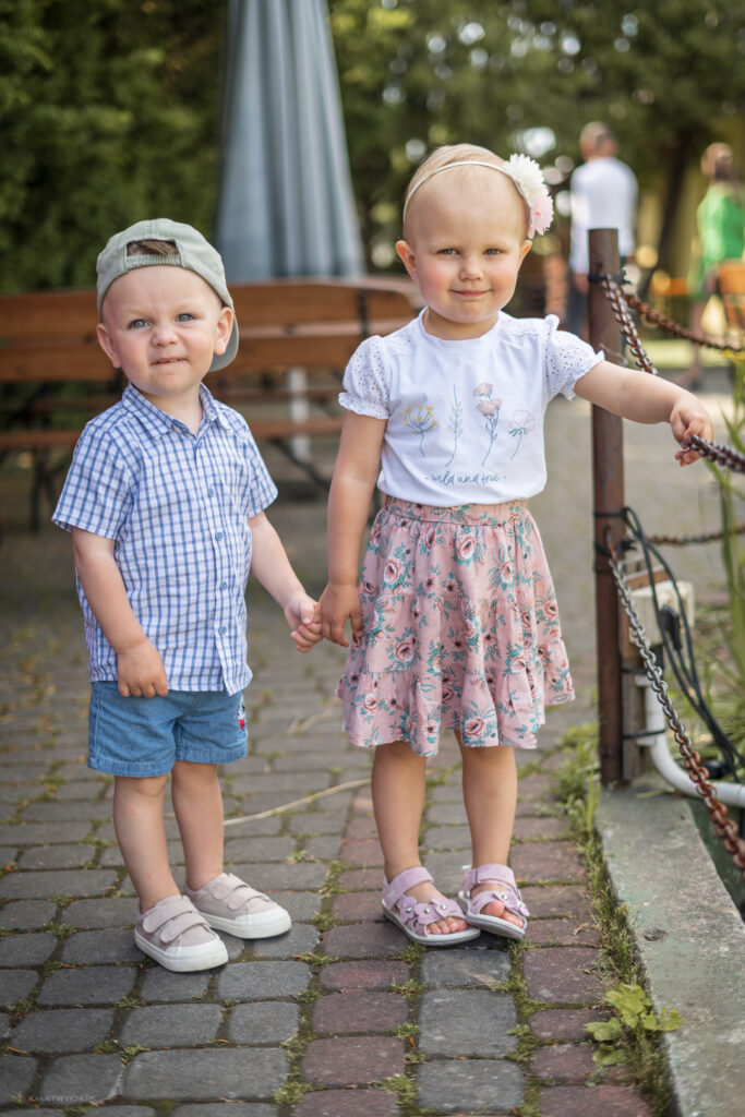 Dwoje dzieci trzymających się za ręce na brukowanej ścieżce przy barierce