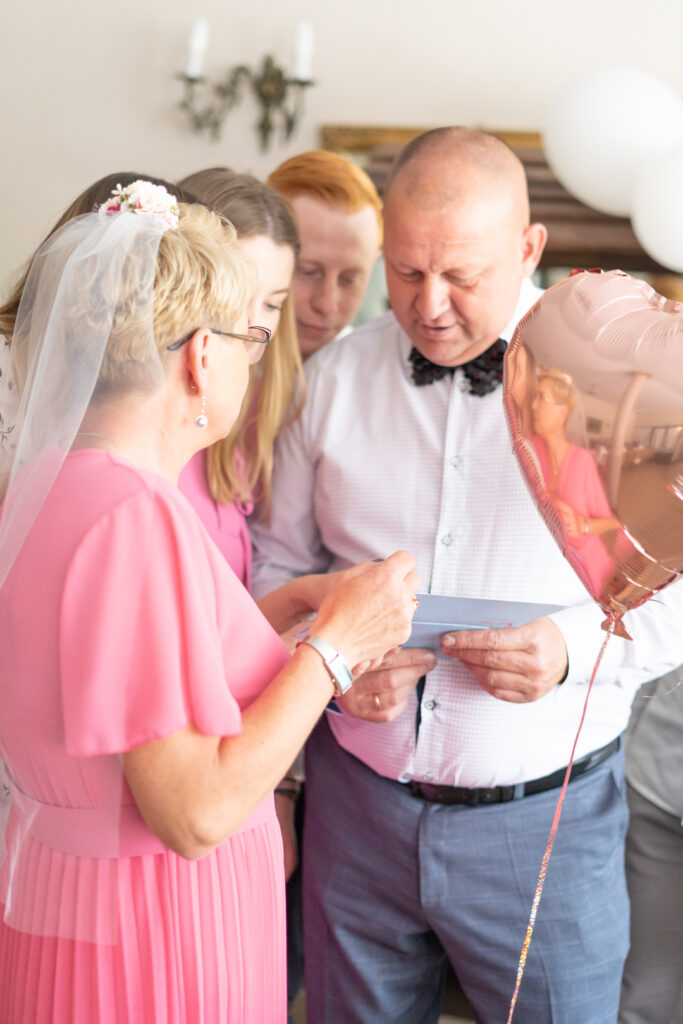 Kobieta w różowej sukni i mężczyzna z muszką oglądają podarunek wśród bliskich