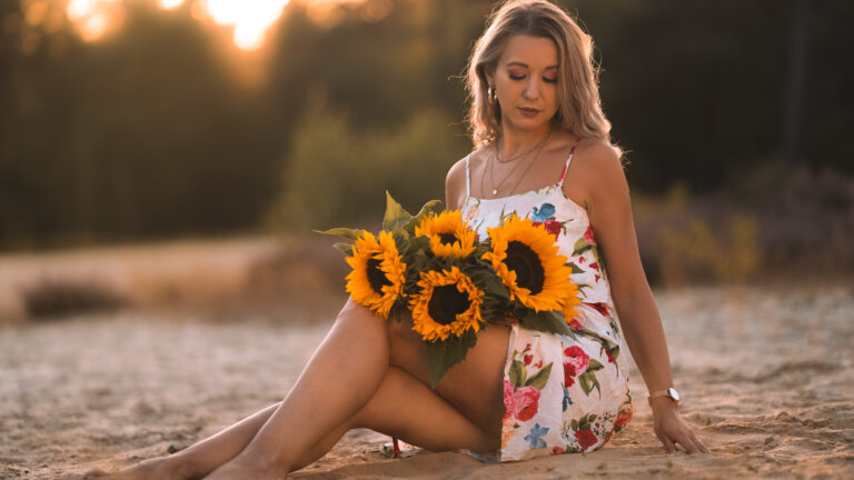kobieta siedzi na piasku w sukience z kwiatami słonecznika